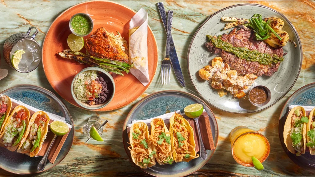 Overhead view of roasted meat, steak, tacos, and drinks on marble.