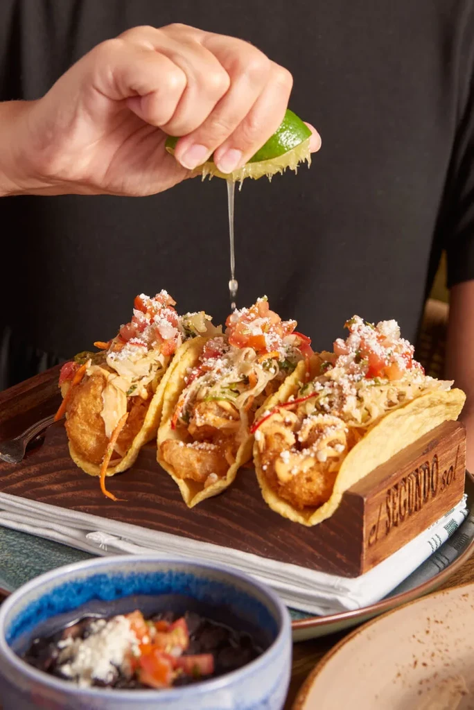 Hand squeezing lime over three fried seafood tacos in a wooden holder.