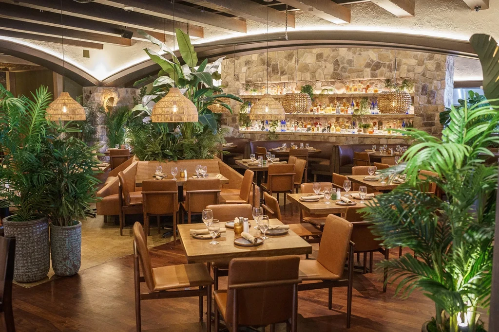 Empty restaurant interior with set wooden tables, a bar, and tropical plants.