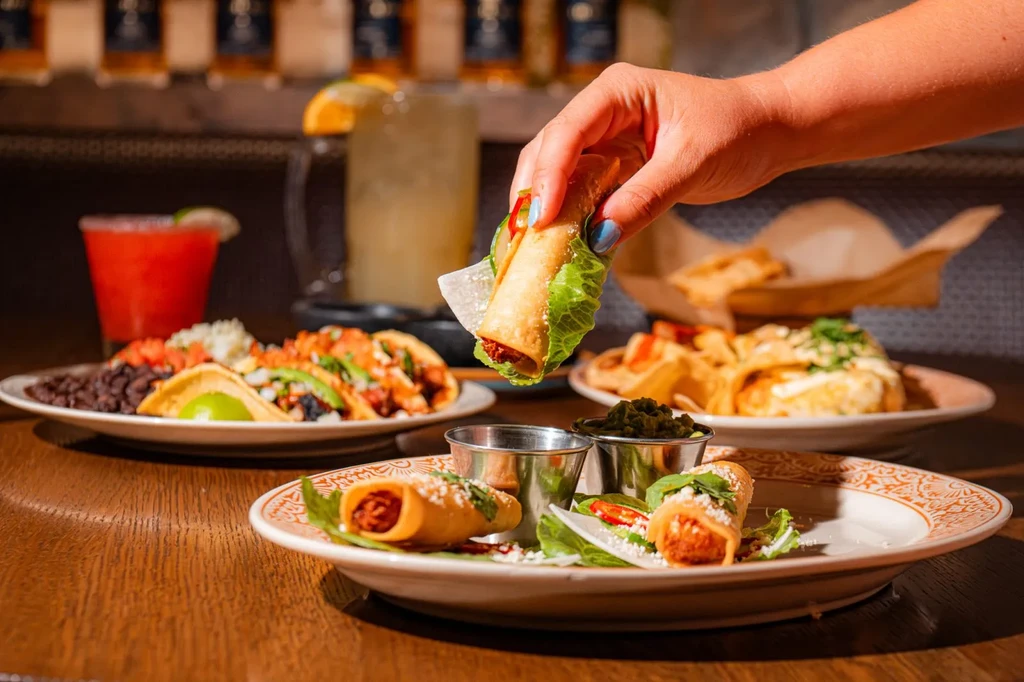 Hand holds a fried taquito over a table of Mexican food.