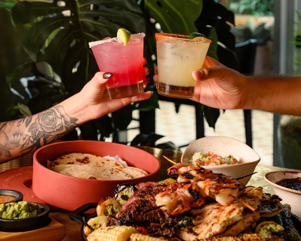Hands with floral tattoo toast margaritas above a grilled food spread.