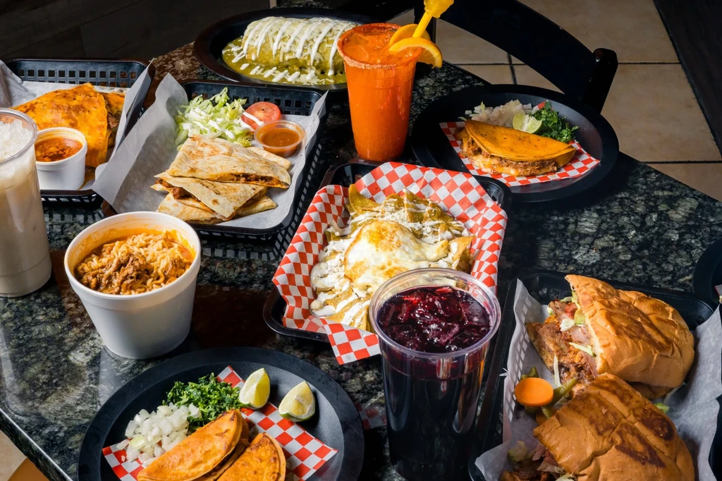 Table spread with diverse Mexican food: tacos, burritos, chilaquiles, torta, drinks.