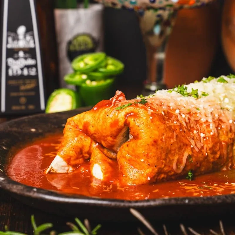 A generously sauced burrito with cheese and fresh herbs on a dark plate.