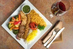 Plated Middle Eastern meal: grilled meat skewers, two rice types, vegetables, sauce.