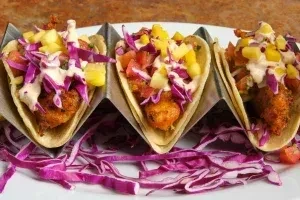 Three fried fish tacos with cabbage, tomatoes, pineapple in a holder.