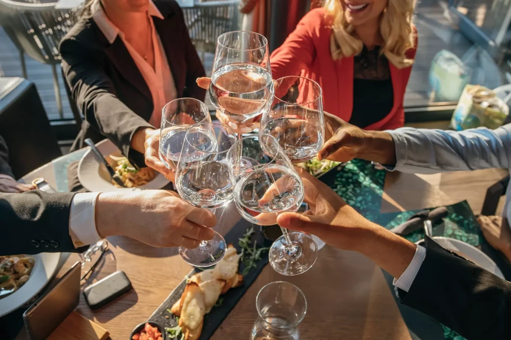 Diverse hands clink water-filled wine glasses over a wooden table.