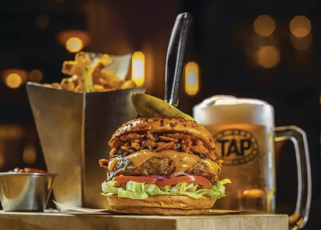 Gourmet burger with pickle spear, fries, dipping sauce, and beer.