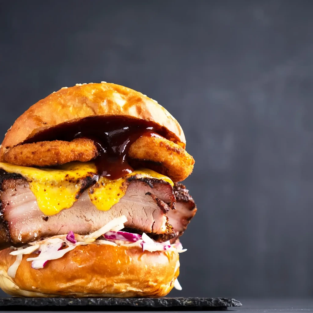Gourmet burger with sesame bun, onion rings, slow-cooked meat, and coleslaw.