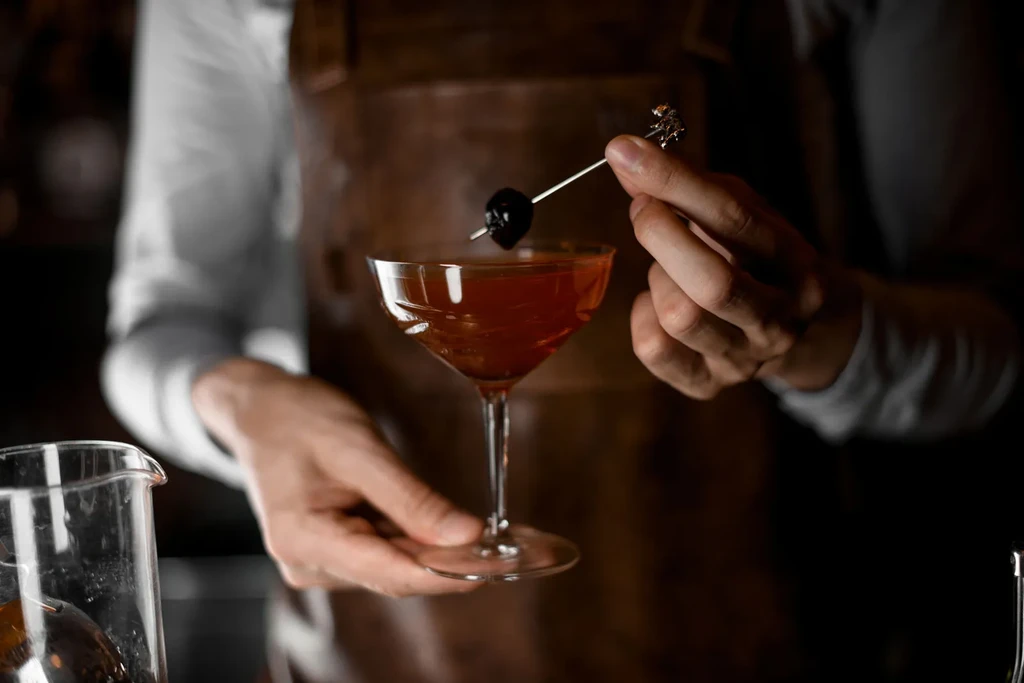 Bartender's hands placing a cherry skewer into an amber cocktail.