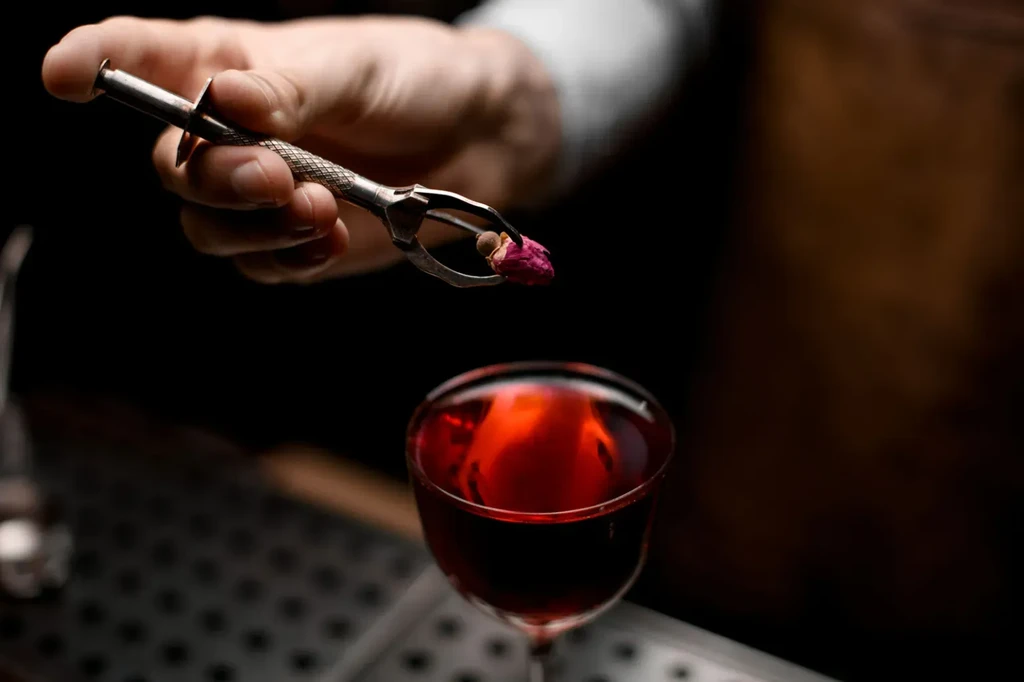 Bartender's hand places a dried rosebud into a red coupe cocktail.