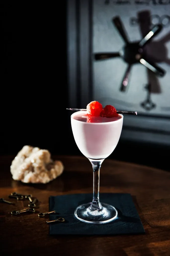 Pink raspberry cocktail, brass keys, with a safe door background.
