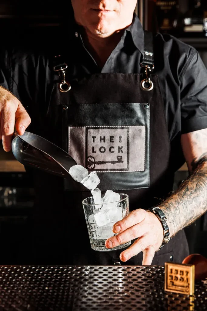 Bartender in 'THE LOCK' apron scoops ice into a glass.