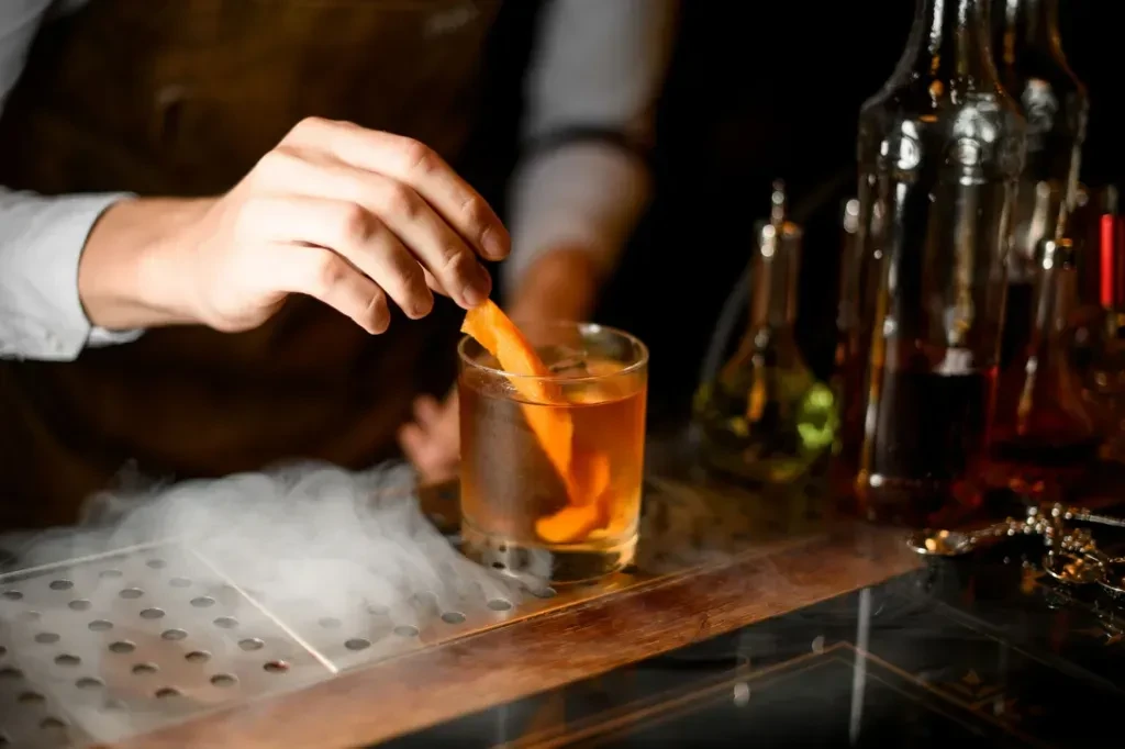 Bartender adds orange peel to amber cocktail with billowing smoke.