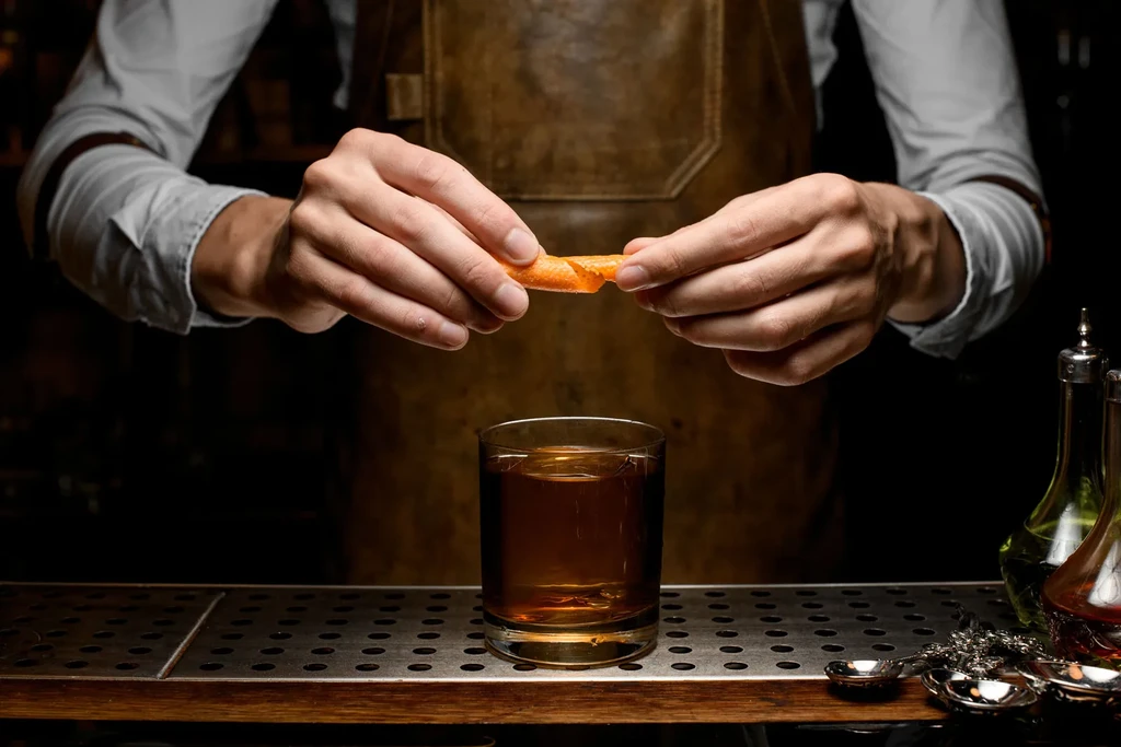Hands in an apron express orange peel over an amber drink with ice.