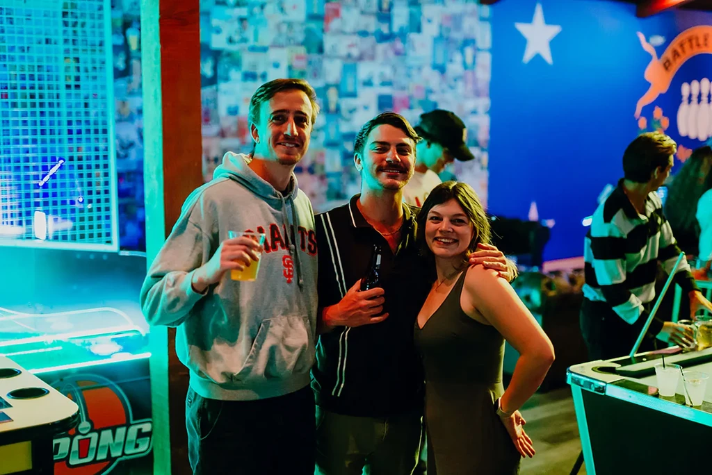 Two men and a woman smile at a neon-lit bar with games.
