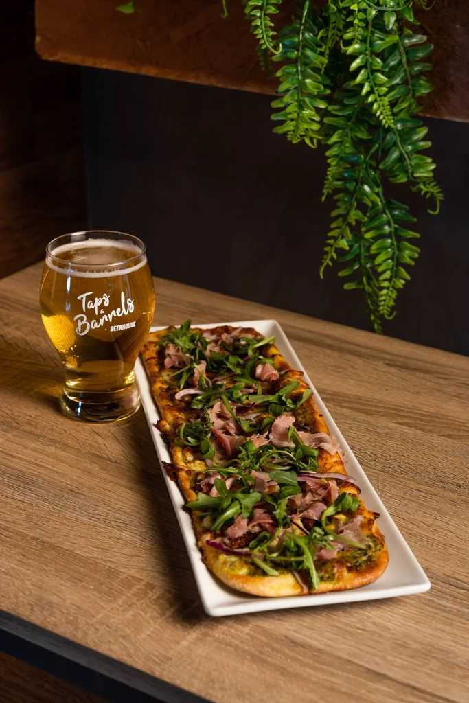 Long flatbread with toppings next to a branded Taps & Barrels beer.