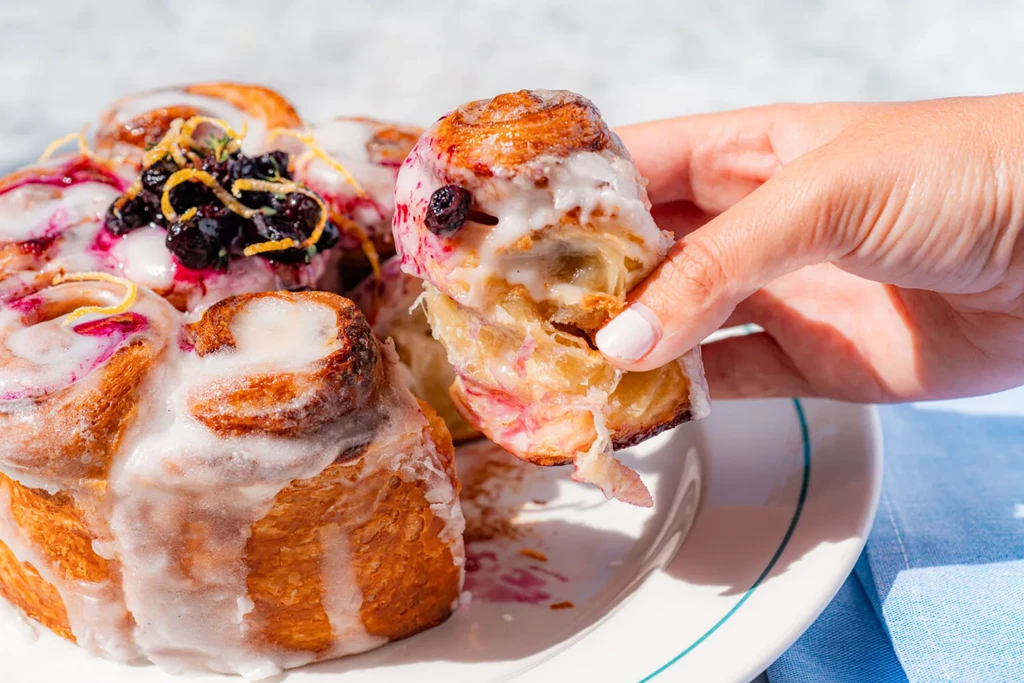 Hand pulls flaky pastry roll topped with icing, blueberries, lemon.
