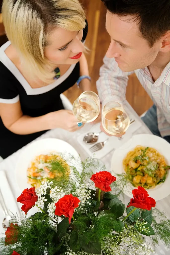 Couple toasts with wine at restaurant with red rose centerpiece.