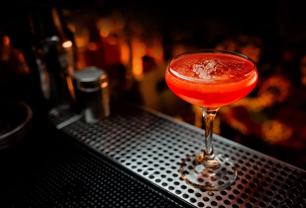 Vibrant red cocktail with gold flakes in coupe glass on bar.