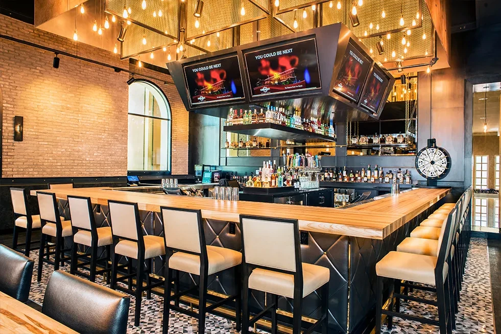Empty, modern L-shaped bar with stools, liquor, beer taps, and TVs.
