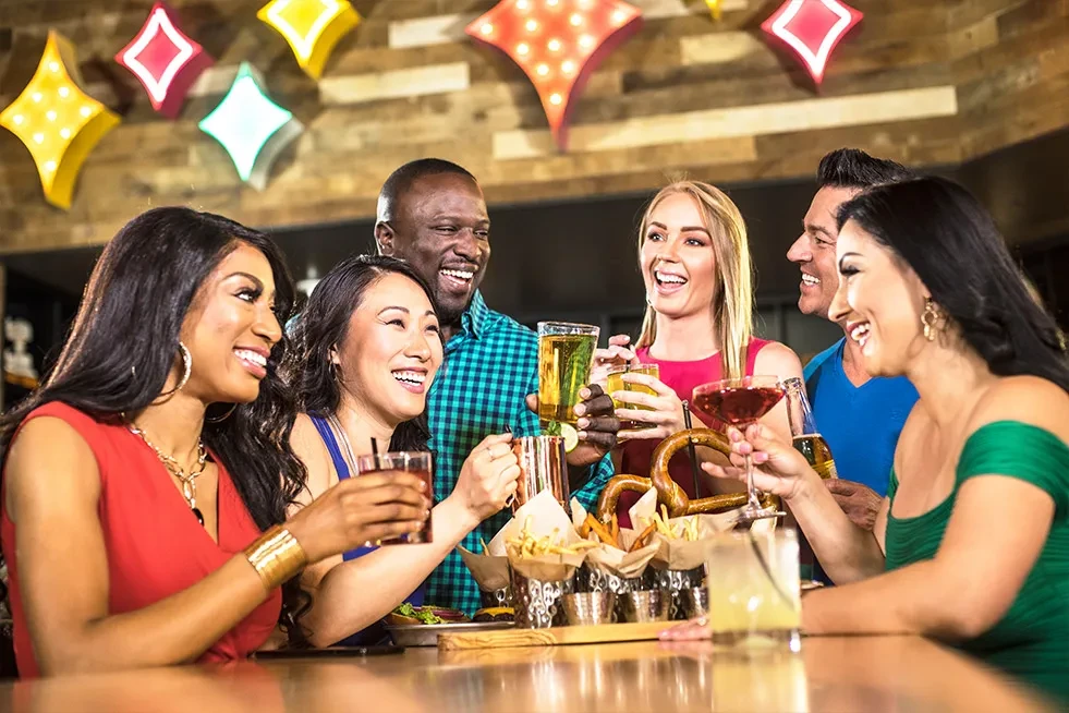 Six diverse people toast drinks and eat snacks at a lively bar.