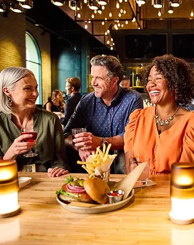 Three people smiling, enjoying burgers, fries, and drinks at a bar.