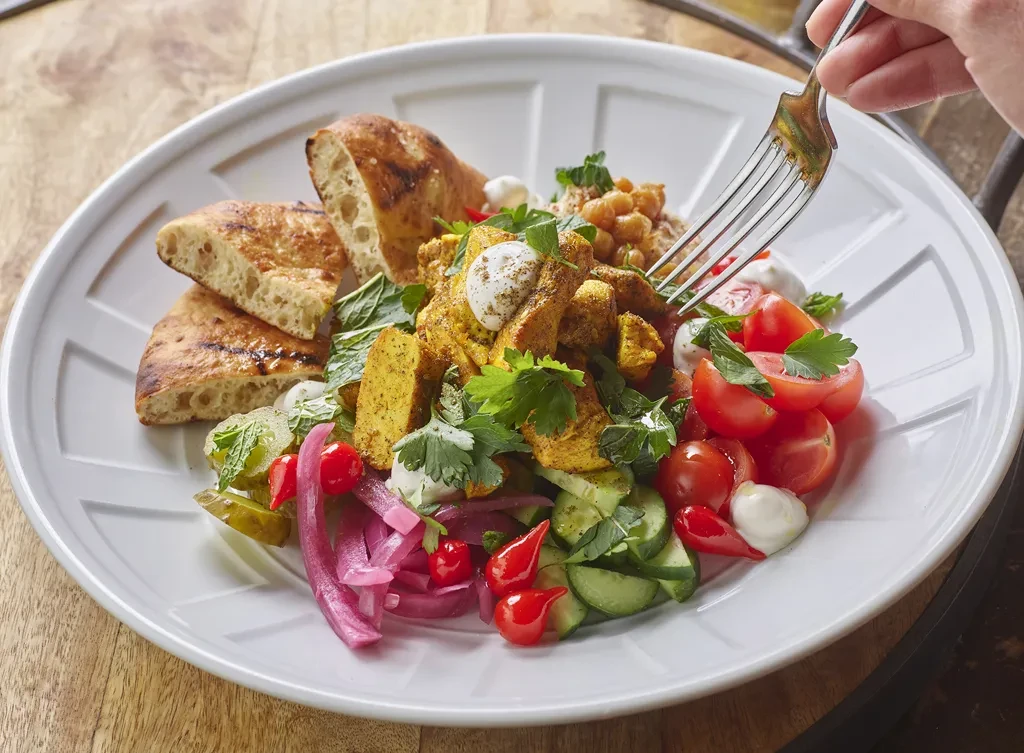 Hand eating vibrant Mediterranean bowl with grilled meat, vegetables, and flatbread.