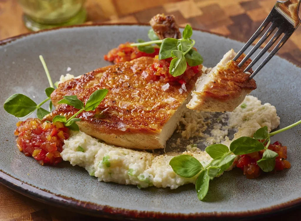Fork lifting crispy pan-seared chicken, puree, red relish, pea shoots.