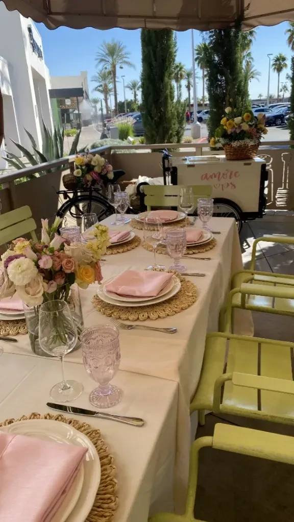 Elegant outdoor dining table with pink flowers, bicycle, and sunny background.