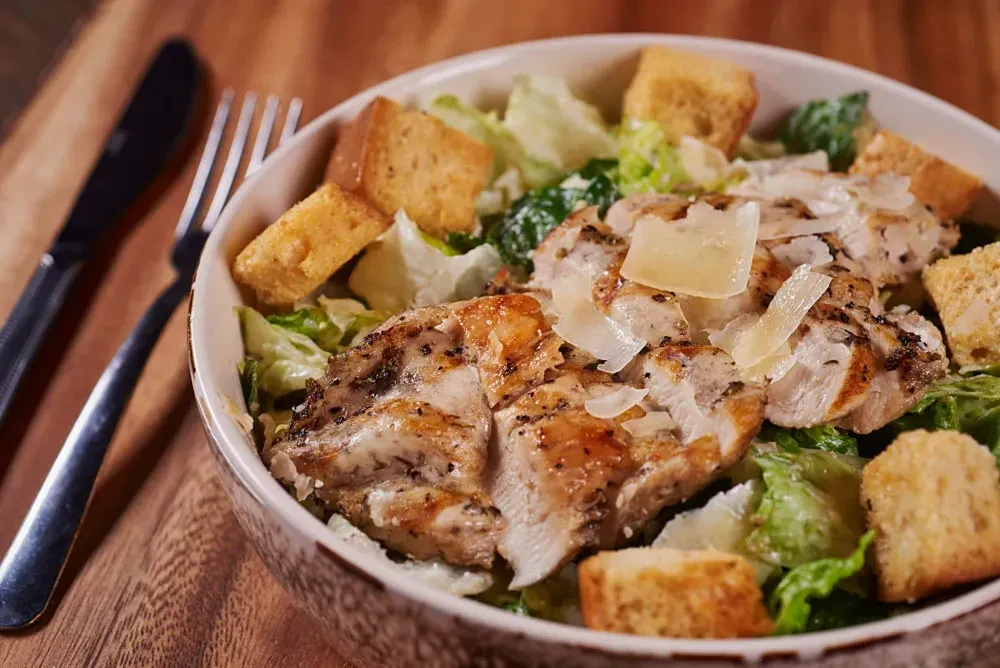 Bowl of Caesar salad with grilled chicken, Parmesan, on rustic table.