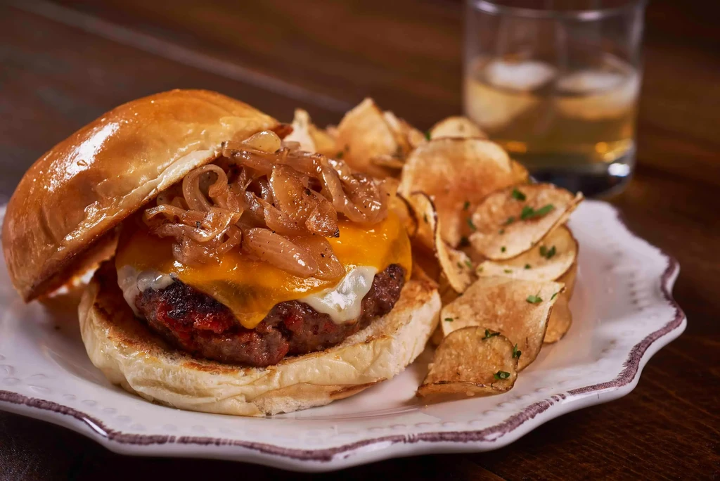 Gourmet burger, herbed potato chips, and amber drink on white plate.