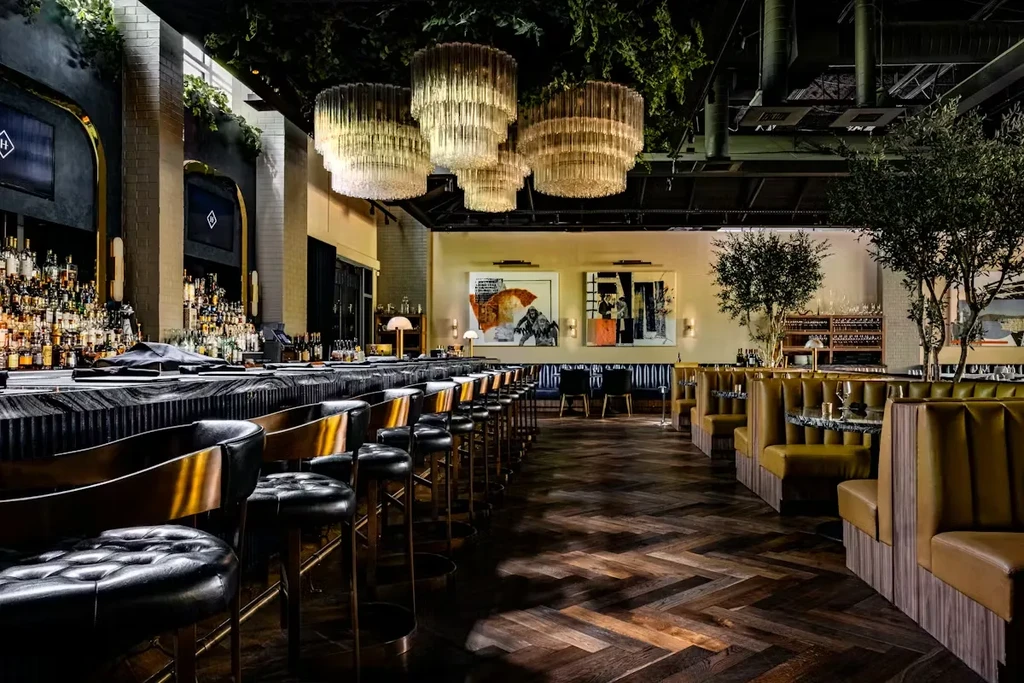 Restaurant interior with a bar, leather stools, chandeliers, and booth seating.
