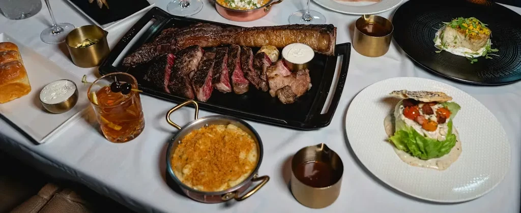 A dining table covered with a diverse spread of steak, pasta, salad, drinks.
