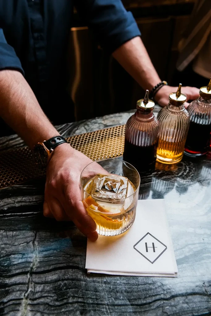 Hand holds amber cocktail with custom ice, orange peel, H-logo coaster.