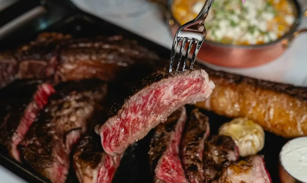 Fork lifting marbled, medium-rare steak slice from dark platter.