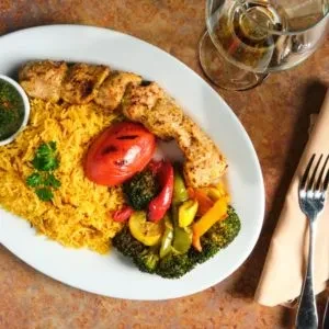 Plated meal: grilled chicken skewers, yellow rice, roasted vegetables, green sauce.