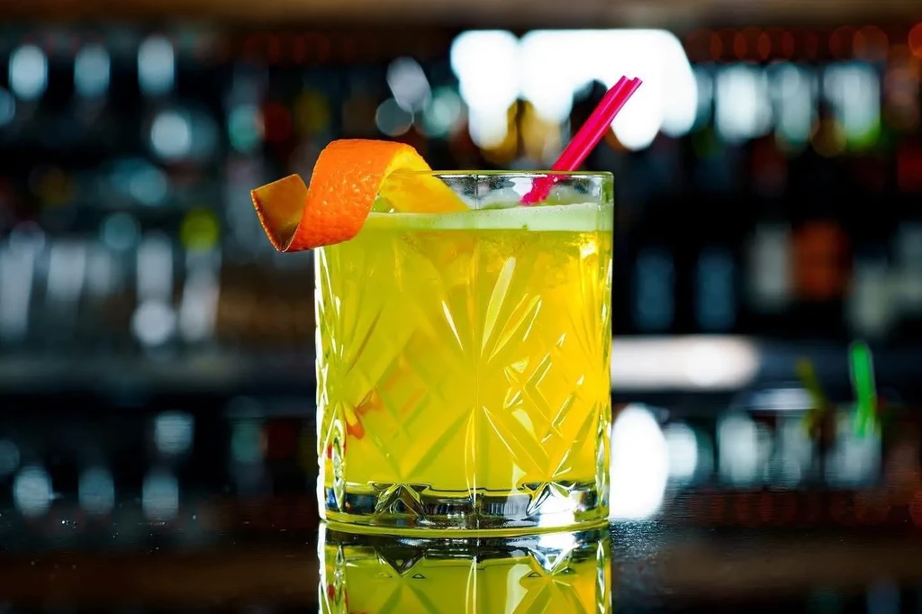 Yellow cocktail in a diamond-cut glass with orange peel and pink straw.