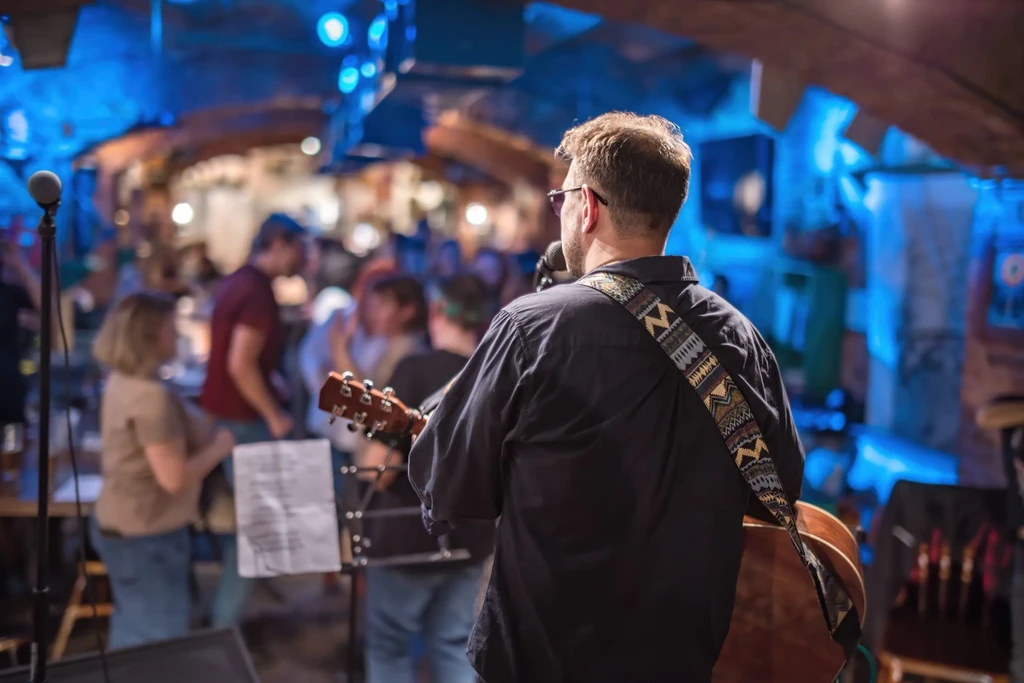 Back of musician playing acoustic guitar in blue-lit bar.