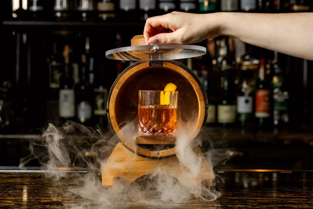 Cocktail in wooden barrel with rising smoke; hand holding lid.