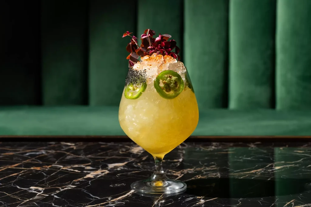 Yellow cocktail with jalapeño slices, spiced rim, and purple herbs.