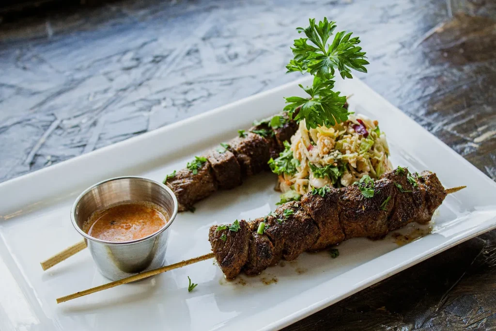Two seasoned meat skewers, salad, and dipping sauce on a white plate.
