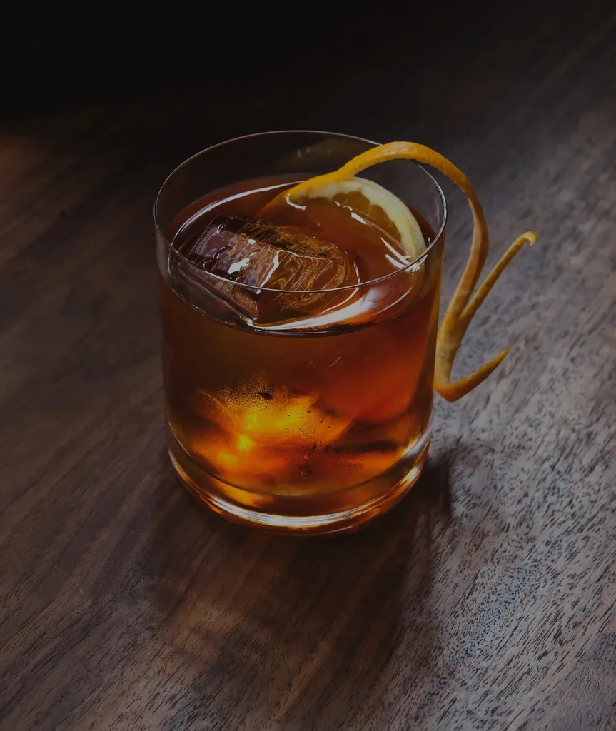 Amber cocktail with large ice, citrus garnish, on dark wood surface.
