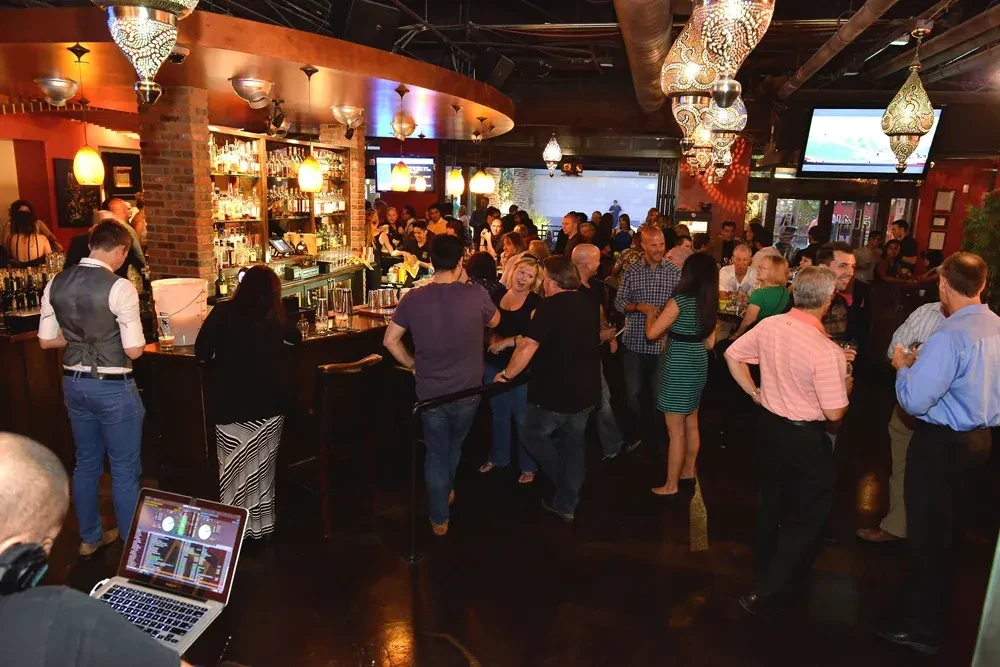 Crowded bar interior with a DJ, ornate lights, and industrial decor.
