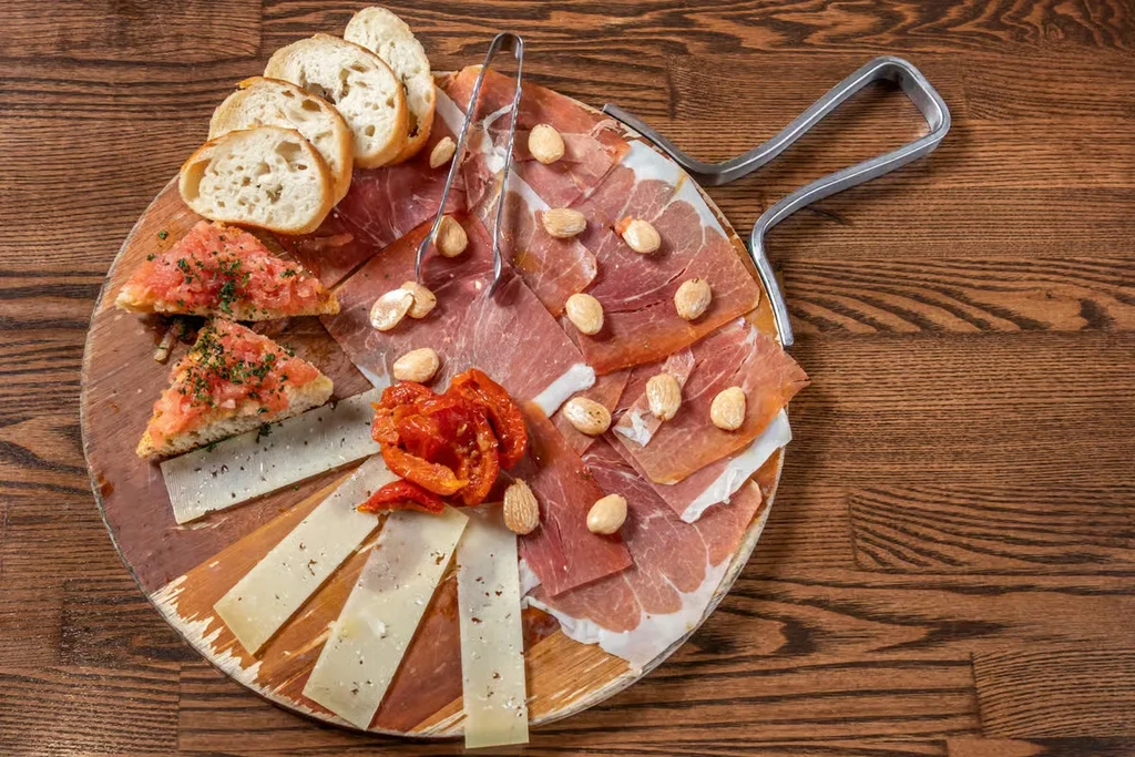 Rustic charcuterie board with meats, cheese, bread, tomatoes, nuts, and tongs.