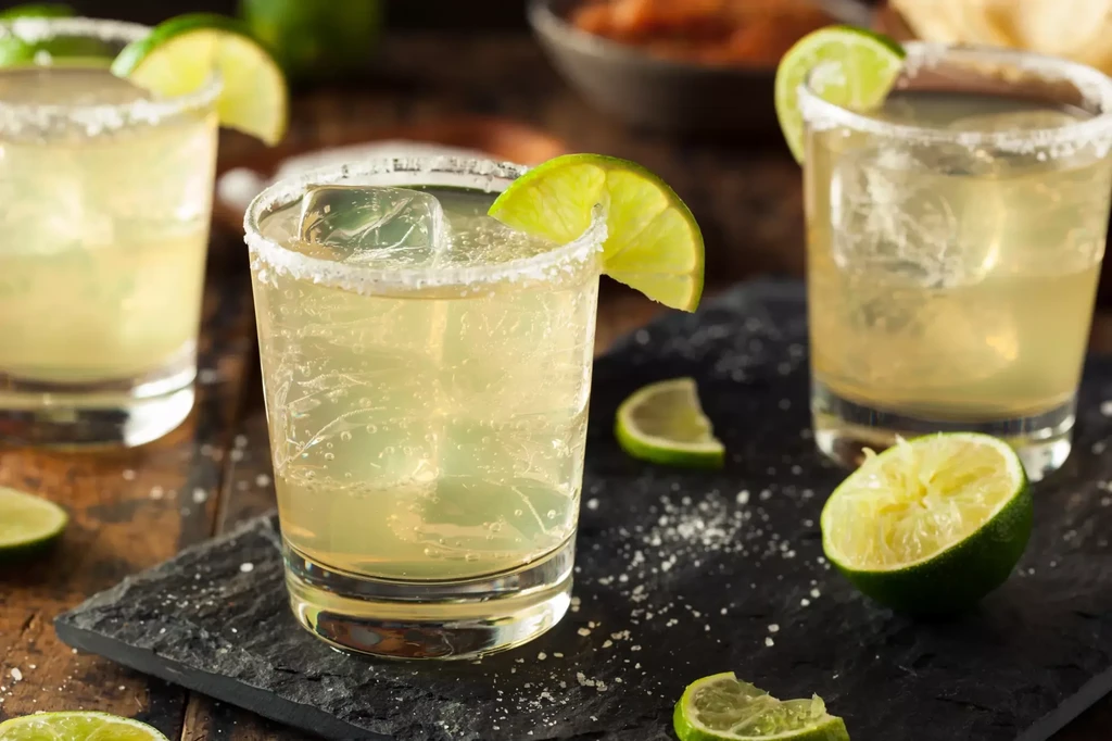 Three lime cocktails with salt rims and wedges on a dark surface.