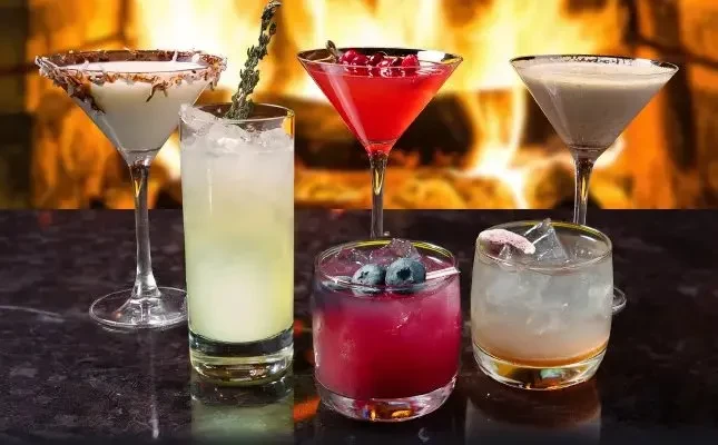 Five colorful cocktails on a dark, reflective surface, fireplace glow.