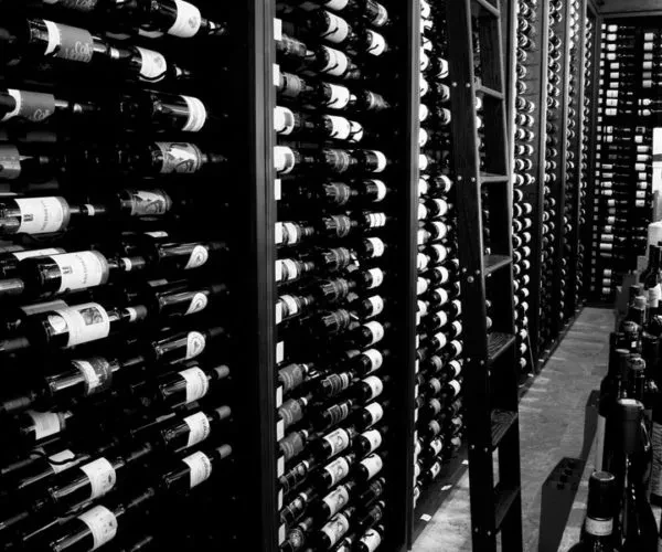 Black and white photo: wine bottles stacked high on shelves in cellar.