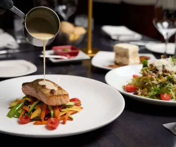 Gloved hand pours sauce over seared fish and julienned vegetables.