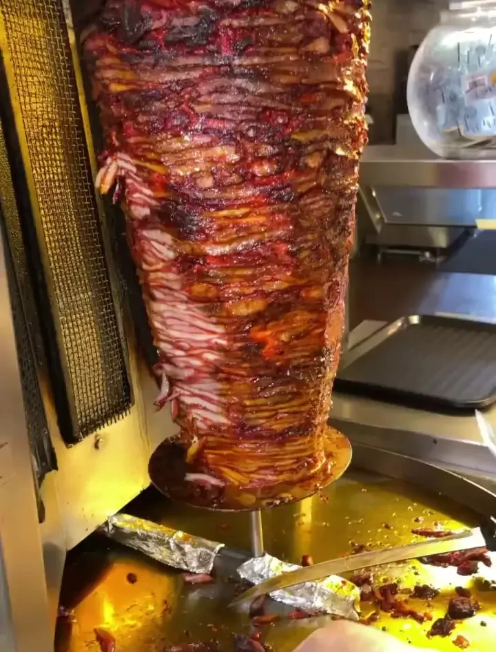 Vertical rotisserie cooking charred al pastor meat, with carved pieces in a tray.