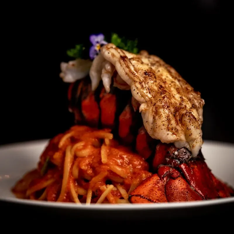 Grilled lobster tail on pasta with reddish sauce, purple flower, herbs.